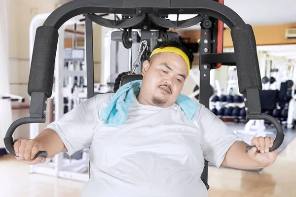 Picture of young obese man looks tired while sitting on the exercise machine. Shot in the gym center