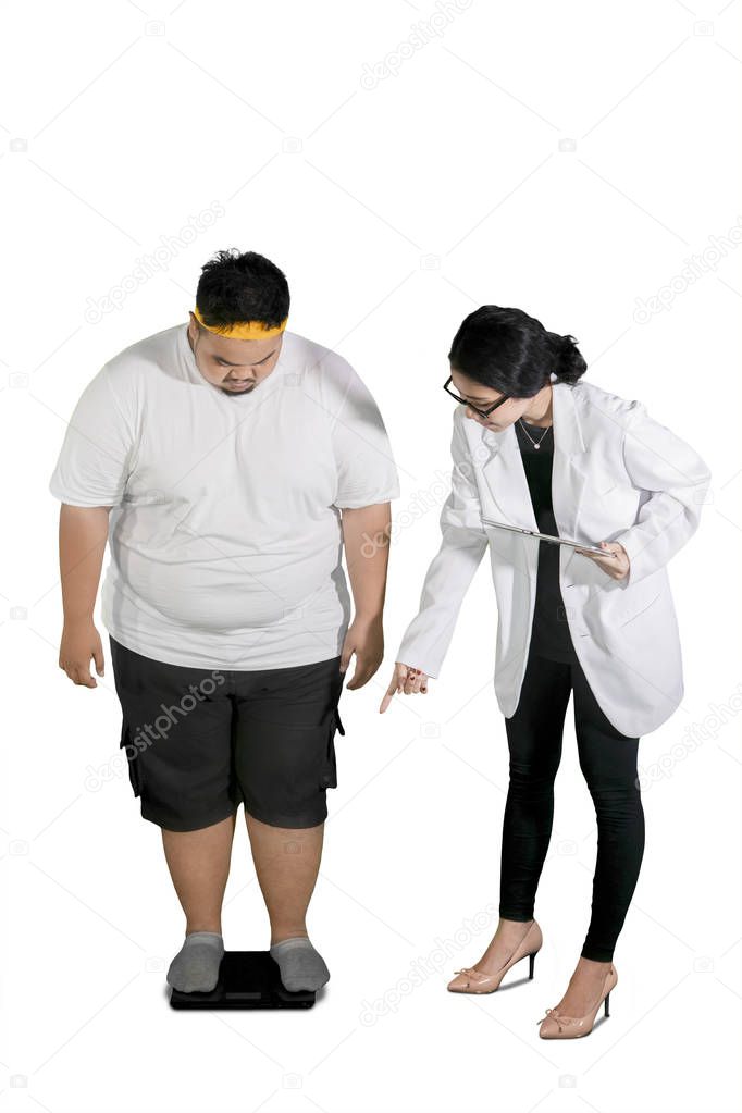Portrait of female doctor examining her fat patient by using scales in the studio, isolated on white background
