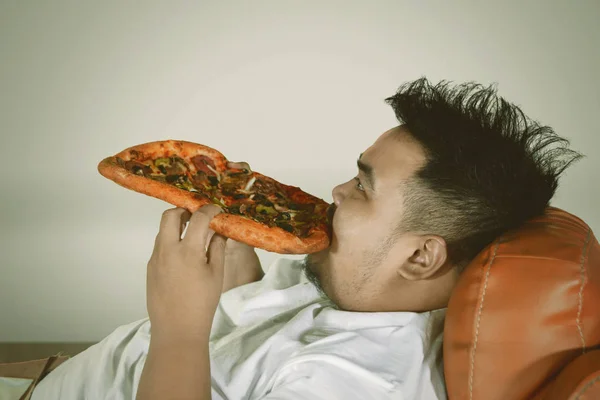 Close up of a young fat man relaxing on the sofa while eating a big pizza