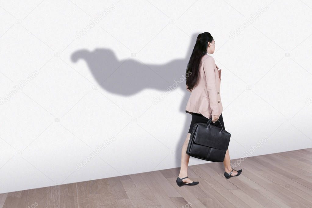 Picture of young businesswoman carrying a suitcase while walking with superhero cape shadow on the wall