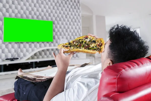 Young fat man eats pizza in front of a television