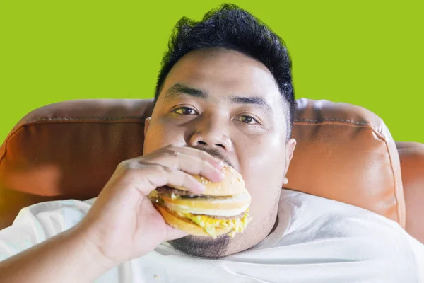 Voracious obese man eating a burger on the sofa