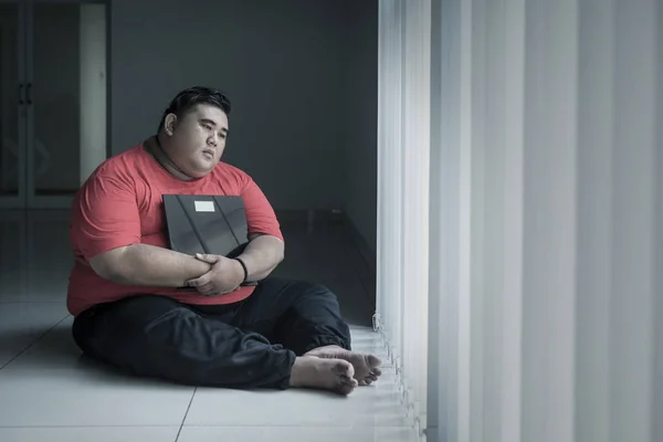 Olycklig fet man som håller en skala nära fönstret — Stockfoto