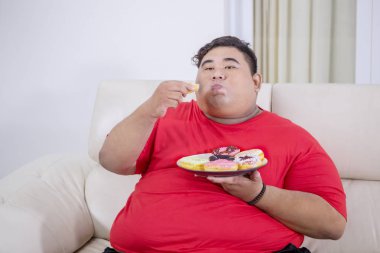 Açgözlü obez adam kanepede çörek bir tabak yiyor
