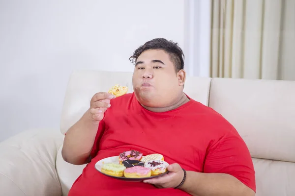 Tlustý muž požívající talíř koblihy na gauči — Stock fotografie