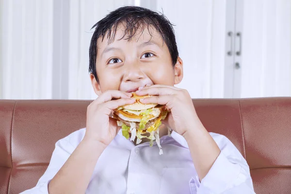 Głodny chłopiec jedzący hamburgera w domu. — Zdjęcie stockowe