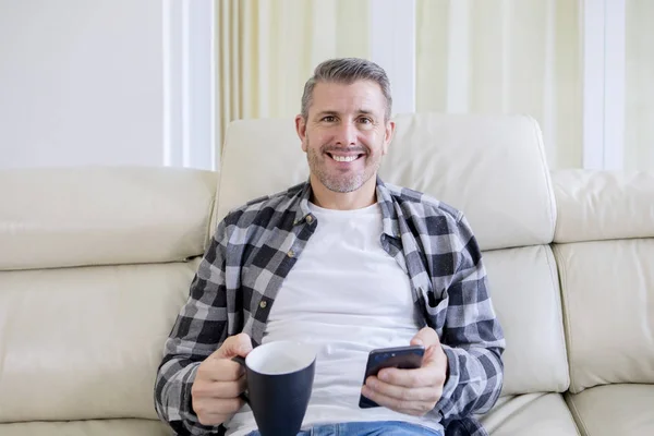 Un homme souriant tient une tasse de thé et un téléphone sur le canapé — Photo