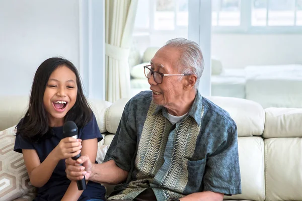 Happy little girl and her grandfather singing together with a microphone while sitting on the sofa in the living room at home