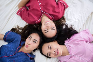 Top down view of three teenage girls smiling at the camera while lying together on the bed and having sleepover party at home clipart