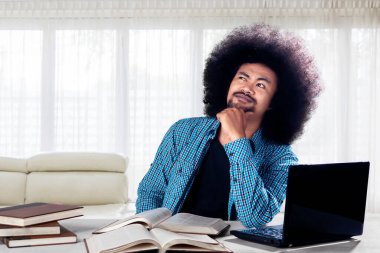 Afrikalı erkek öğrenci evde dizüstü bilgisayar ve kitaplarla çalışırken bir fikir düşünüyor.