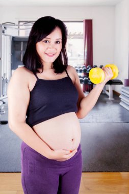 Hamile kadın halterle egzersiz yaparken ve spor salonunda dikilirken mutlu görünüyor.