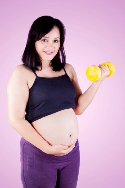 Genç hamile kadın halterle egzersiz yaparken ve stüdyoda ayakta dururken mutlu görünüyor.