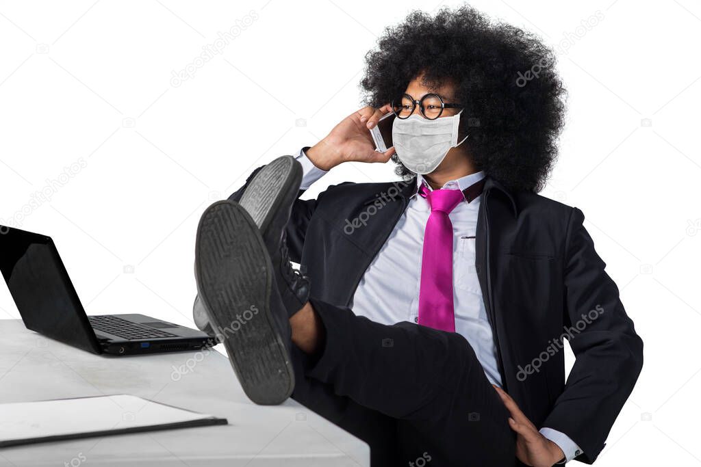 Afro businessman talking on a cellphone while sitting with laptop and his foot on the table in the studio, isolated on white background