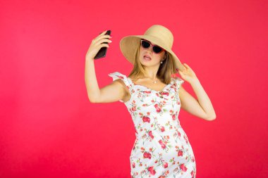 Göz kamaştırıcı beyaz kadın cep telefonuyla selfie çekiyor. Stüdyoda güneş gözlüğü ve yaz elbisesi takıyor. Kırmızı arka planda.