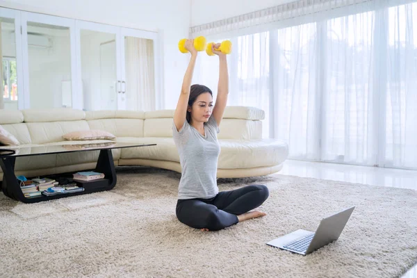 アジアの女性運動とともにダンベルとともにビデオを見ながらラップトップコンピュータ上で自宅で自己検疫を行う — ストック写真