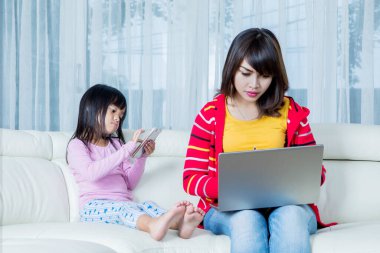 Genç anne evde dizüstü bilgisayarla çalışırken meşgul görünüyor ve kanepede akıllı telefon kullanarak kızını görmezden geliyor.