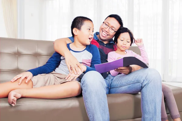 快乐的父亲和他的两个孩子坐在沙发上看书 在客厅里放松 在家里中弹 — 图库照片