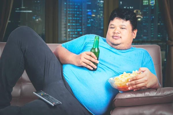 Obesity man eating chips and drink a beer in the living room while watching TV and lying on the sofa. Shot in the apartment