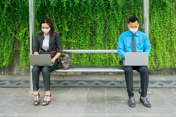 两个商人一边戴面具一边用笔记本电脑 一边在巴士站以新的正常生活方式进行社交活动 — 图库照片