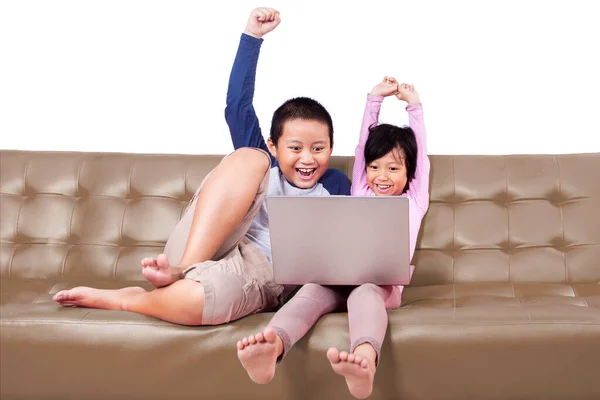 两个小孩一起欢呼和举手 一边在笔记本电脑上玩游戏 一边坐在沙发上 在有白色背景的独立演播室里被枪杀 — 图库照片