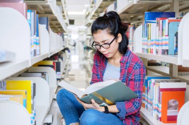 Kütüphane koridorunda oturmuş kitap okuyan bir üniversite öğrencisi.