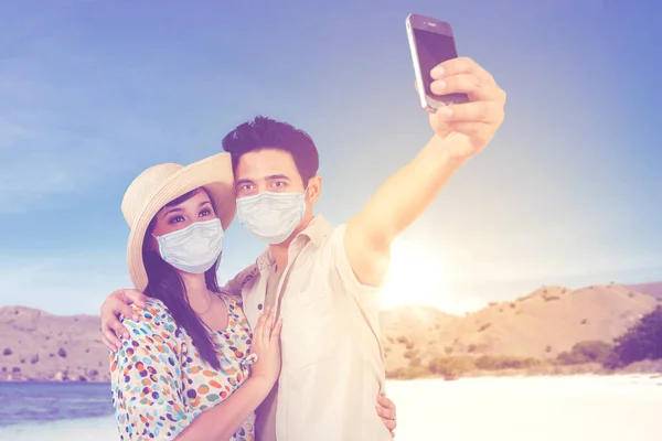 这对浪漫夫妇戴着面具 一边用智能手机拍照 一边站在沙滩上晒太阳 — 图库照片