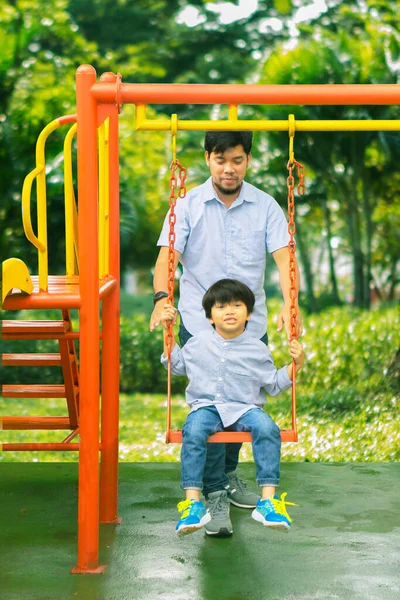 可爱的小男孩坐在秋千上 和他的父亲一起在操场上玩耍 在公园里享受着闲暇时光 — 图库照片