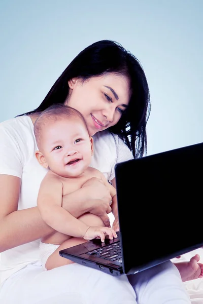 幸せな母親保持彼女の赤ちゃんながら使用してノートパソコンと座ってスタジオでグレーの色の背景 — ストック写真
