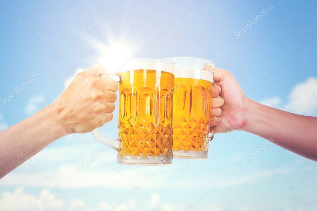 Hands of two people holding a glass of fresh beer while making toasts under blue sky. Shot at outdoors