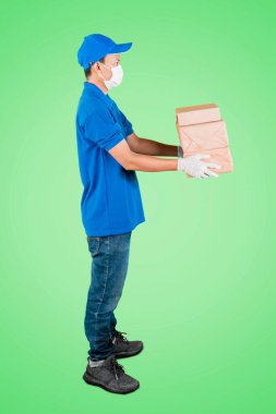 Yüz maskesi ve eldiven takmış bir erkek kuryenin bir yığın paketi teslim ederken ve yeşil ekran arka planıyla stüdyoda dikilirken yan görüntüsü