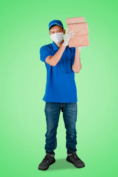 パッケージのスタックを運ぶと緑の画面の背景を持つスタジオに立っている間 顔のマスクと手袋を身に着けている男性の宅配便の完全な長さビュー — ストック写真