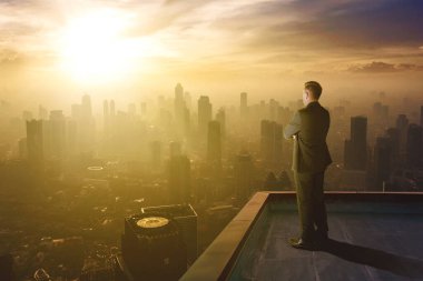 Erkek yönetici gün batımında gökdelenleri olan sisli şehre bakarken binanın çatısında duruyor.