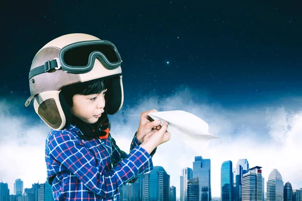 可爱的小男孩戴着头盔 一边玩纸飞机 一边站在有现代城市背景的地方 在室外开枪 — 图库照片
