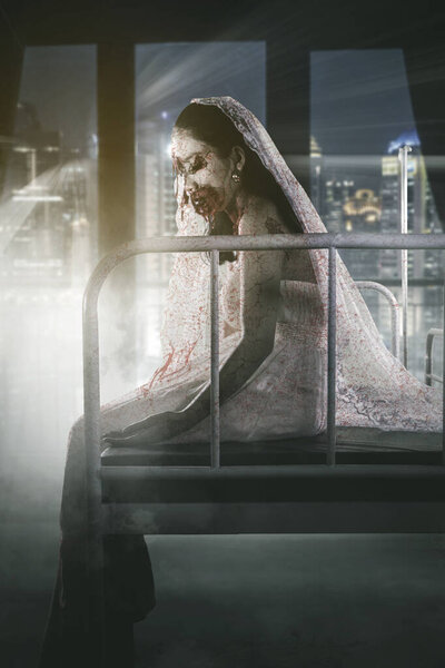 Концепция ужасов Хэллоуина. Ужасная невеста призрак с раненым лицом сидит на кровати с современным видом на город фон на окне