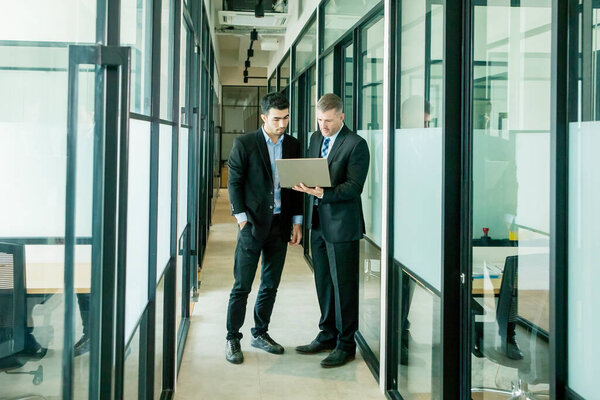 Два мультиэтнических бизнесмена обсуждают в офисном коридоре, стоя и используя ноутбук