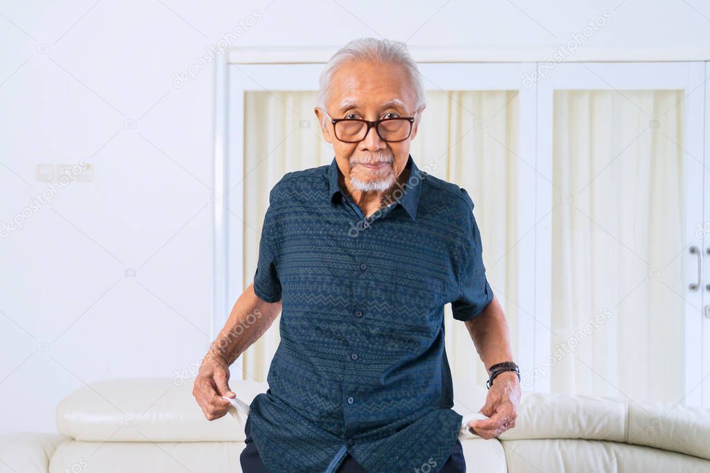 Elderly man showing his empty pocket while standing in the living room at home