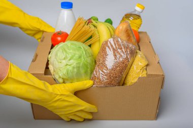 Çeşitli yiyeceklerle birlikte bağış kutusu. İçinde yağ, konserve yiyecek, mısır gevreği ve makarna olan karton bir kutu açın. Sarı eldivenli eller.