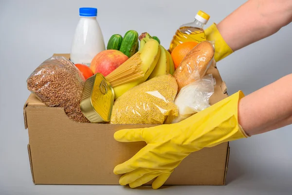 Çeşitli yiyeceklerle birlikte bağış kutusu. İçinde yağ, konserve yiyecek, mısır gevreği ve makarna olan karton bir kutu açın. Sarı eldivenli eller.