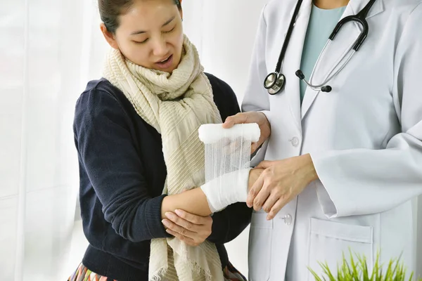 Accidente de mano Lesión, Asiática médico femenino con estetoscopio vendaje mano de paciente en el hospital, Mujer cara de paciente expresar lo doloroso en su muñeca rígida . — Foto de Stock