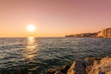 Güzel gün batımı Piran şehir, Slovenya üzerinde. Sihirli güneş Adriyatik Denizi ve Piran sahile ışığı. Yumuşak ışık gökyüzü ve mavi su ön planda. Taş duvara yakın Piran liman.