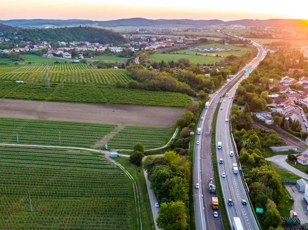 Aerial above view of highway road with cars. Highway leads from bottom to top through the trees and bushes. City panorama with buildings in background. Sunny day, sunset — Stock Photo, Image