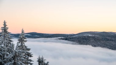 Kış günbatımının tersine karla kaplı tepeler ve bulutların üzerinde yükselen ormanlar, yukarıdan çekilen sıcak akşam ışığı ile parlayan tonlarda gökyüzünü boyuyor..