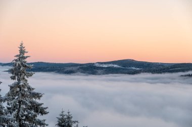 Kış günbatımının tersine karla kaplı tepeler ve bulutların üzerinde yükselen ormanlar, yukarıdan çekilen sıcak akşam ışığı ile parlayan tonlarda gökyüzünü boyuyor..