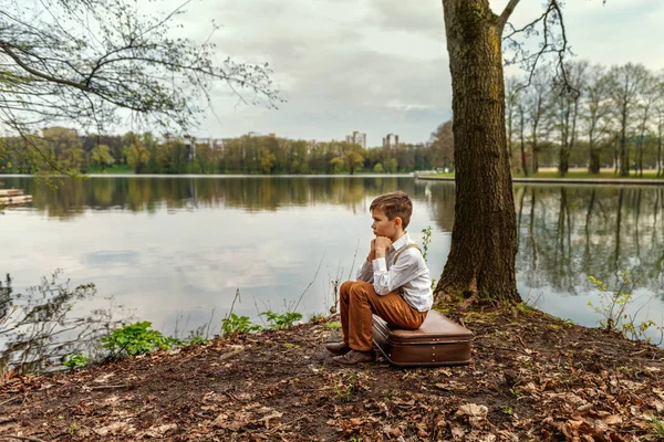 a young nipper guy in retro old-fashioned village clothes sits saddened on an old suitcase propped his head on the bank of a river lake with his fists