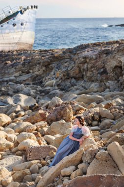 güzel bir bayanı incelikle zarif ve arka plan üzerinde terk edilmiş bir tekne ile bekleyen taş oturur.