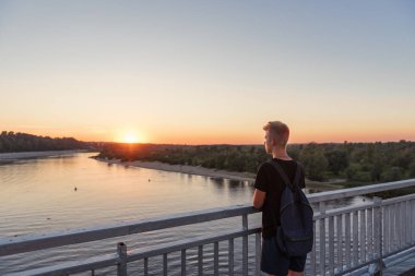 young guy teenager standing on bridge over river water in lifestyle clothes near steel railing and looking afar at sunset clipart