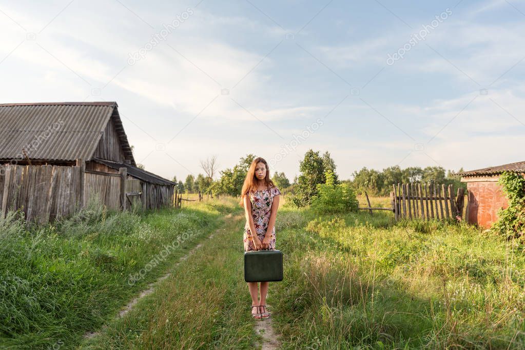 a model girl in a retro dress is holding in her hands a vintage suitcase on an abandoned country road overgrown with grass between the village courtyards and huts