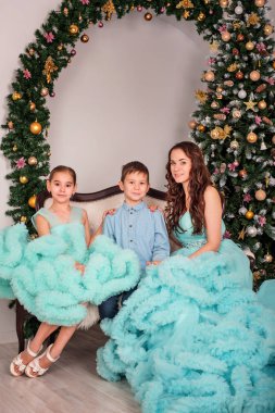 Onun oğlu ve kızı stüdyoda fotoğraf oturumunda zarif mavi elbiseler ile anne Noel ağacının yanında oturur