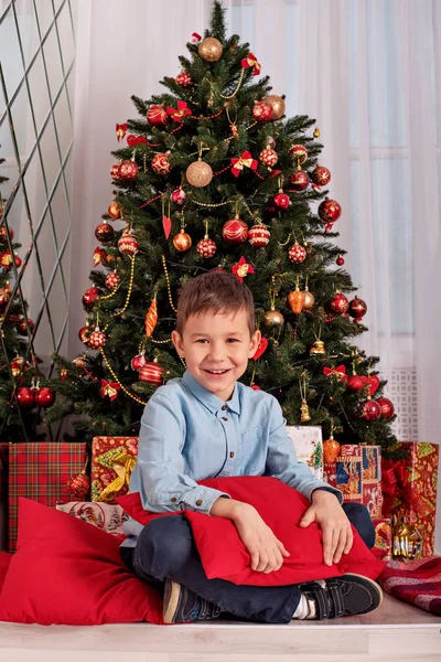 boy sitting near the Christmas tree hugging a cozy pillow on New Year\'s Eve