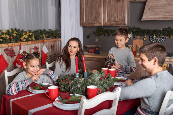 a family of four people in identical sweaters sit at the Christmas table and the boy pours tea in the New Year\'s decorated kitchen